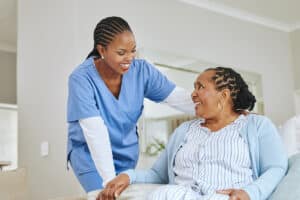 A nurse in blue scrubs warmly smiles at an elderly woman sitting on a couch.