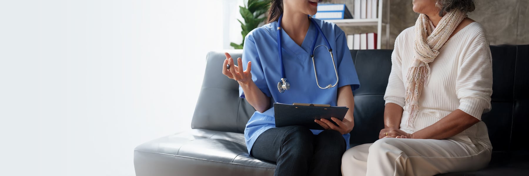 A nurse in blue scrubs and stethoscope talks empathetically with an elderly woman on a couch.