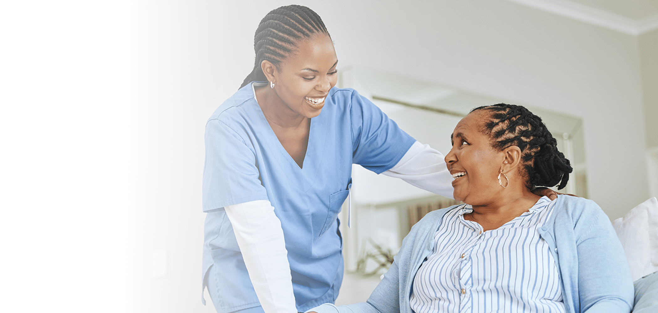 A nurse in blue scrubs warmly smiles at an elderly woman sitting on a couch.